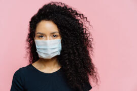 Serious young woman being on self isolation at home, wears protective medical mask, being on quarantine at home, dressed casually, poses against pink background. Coronavirus disease